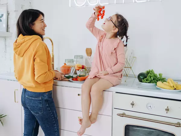 Young girl seated on kitchen counter next to mother looking mindfully at cherry tomato vine.