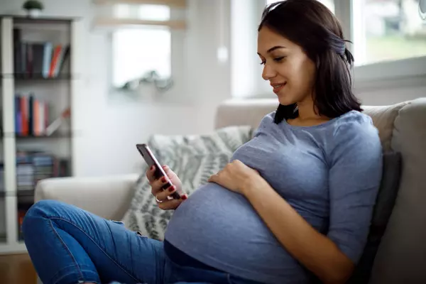 Smiling pregnant woman using cell phone on couch.