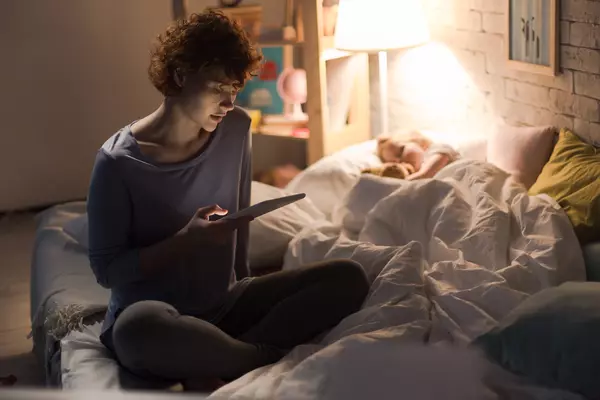 Mother on tablet next to sleeping child in bed.